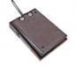 Preview: The image shows a rectangular, flat device with a rough surface texture. It has two push buttons on top and a cable that leads out from the side. The color is dark brown.