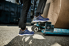 Preview: The image shows a standing man in dark clothing wearing blue safety shoes. He is standing next to a cart with a large, round box on a paved area.