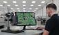 Preview: A man is sitting at a table, looking at a green circuit board with components on a monitor. The room is bright and modern, with additional workstations in the background.