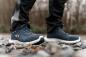 Preview: The image shows two feet in blue, sturdy shoes standing on a pebbly ground. The surface is uneven, surrounded by a blurry, natural background.