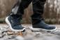 Preview: The image shows a person standing on a rocky surface. They are wearing dark blue, sporty shoes with light soles and are dressed in dark pants. The background is blurred.