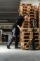 Preview: A man leans over a stack of wooden pallets. He is wearing a black jacket and gray work pants. Next to him is a handcart with a large roll. Bright hall in the background.