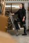 Preview: A man with curly hair stands on a ladder in a warehouse. He is wearing black work clothes and lifts one leg onto a rung while looking upwards. Boxes surround him.