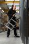 Preview: A man in black work clothes stands in a warehouse holding an aluminum ladder. He looks up at the shelves to reach something. The environment appears industrial and well-organized.