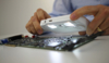 Preview: A person holds a light-emitting magnifying glass over a green circuit board. The hands are visible, and the device displays components such as chips and holes for connections.