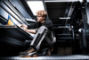 Preview: A young man in work clothes is sitting on a metal floor studying documents. He has curly, light hair and is wearing shoes with orange accents. Behind him is an industrial space.