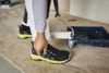 Preview: The image shows a craftsman holding a paint roller in his hand. His shoes are black with yellow accents. He stands next to a paint container on a wooden floor.