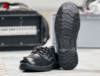 Preview: The image shows a black safety shoe from Sievi. The shoe features simple lacing and a textured, non-slip sole with a distinct tread. In the background, tools are blurred.