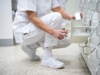 Preview: A person in white clothing and white shoes is sitting on the floor, reaching into a shelf to take something out. The floor is light and the surroundings appear clean and tidy.