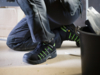 Preview: The image shows a person squatting. They are wearing black work shoes with green accents. The background is blurry, featuring a bucket and boxes nearby.