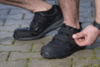 Preview: The image shows a man tightening a black Velcro shoe with one hand. He is standing on a cobblestone path with green moss between the stones.