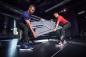 Preview: Two men are lifting a table together in a modern room. One is wearing a blue shirt and sneakers, the other a red shirt and black pants. They are working intently.