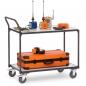 Preview: A gray cart on wheels with two levels. On the top, there are various devices in blue and orange, and below is a large, orange suitcase with a black border. The cart has a handle.