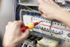 Preview: A person is working on a control cabinet with electrical components. In one hand, they are holding a screwdriver. Several fuses and cables are visible. A technical environment.