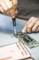 Preview: A hand holds a screwdriver and repairs a green circuit board. On the table, more tools are placed in a pink container fitted with slots for various screwdrivers.