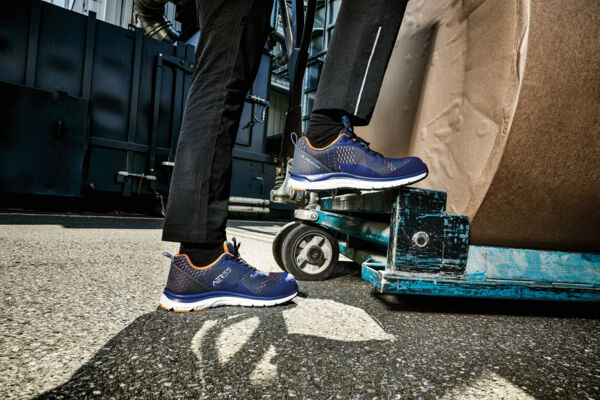 The image shows a standing man in dark clothing wearing blue safety shoes. He is standing next to a cart with a large, round box on a paved area.