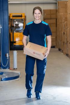 In the picture, there is a woman in blue work attire carrying a cardboard box. She is smiling and is in a warehouse with many boxes in the background.