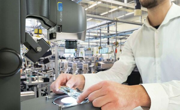 A person in a white shirt is working at a table, examining a circuit board under a microscope. In the background, machines can be seen in a bright factory.