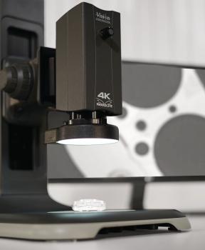 The image shows a microscope with a black lens, aimed at a small, transparent sample. In the background, there is a graphical representation reminiscent of film.