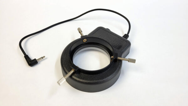 The image shows a round device with a black casing and a central, hollow ring. It has two small levers and a cable that can be connected to a power source.