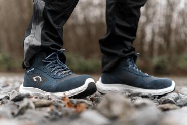 The image shows two feet in blue, sturdy shoes standing on a pebbly ground. The surface is uneven, surrounded by a blurry, natural background.