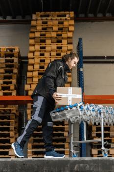 A man in work clothes is lifting a box in a warehouse. In the background, wooden pallets are stacked. He is standing next to a cart with several boxes.