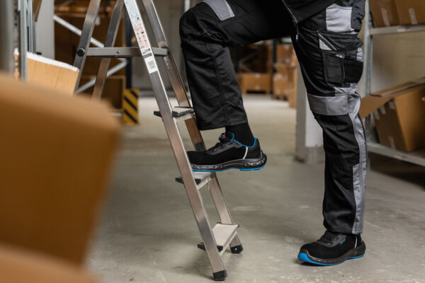 In the picture, you see a man standing on a ladder. He is wearing black work clothes with gray and blue accents. In the background, there are boxes in a warehouse.