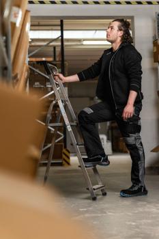 A man with curly hair stands on a ladder in a warehouse. He is wearing black work clothes and lifts one leg onto a rung while looking upwards. Boxes surround him.