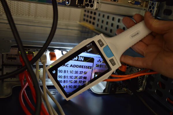 The image shows a handheld device displaying various MAC addresses. It is situated in a technical environment surrounded by numerous cables and an open case.