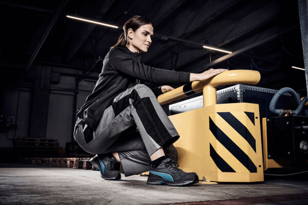 A sportily dressed woman is kneeling at a yellow device in an industrial hall. She is wearing gray work clothes and looking forward intently. The environment is modern and functional.