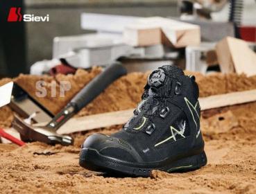 The image shows a black, sturdy work shoe with striking green accents. It stands on sandy ground, surrounded by tools such as a hammer.