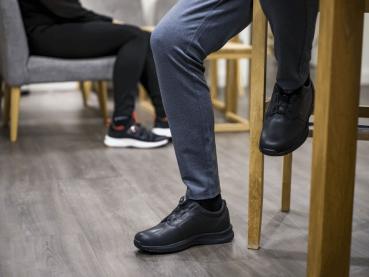 The image shows two people in a café. One person is sitting on a chair with black shoes and gray pants. The other person is in the background, also wearing black sneakers.