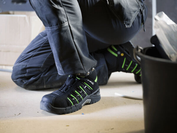 The image shows a person squatting. They are wearing black work shoes with green accents. The background is blurry, featuring a bucket and boxes nearby.