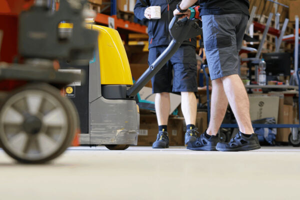 In the image, there are two people wearing shorts operating a pallet jack. One is holding a cup. The floor is light-colored and the surroundings have shelves filled with crates.