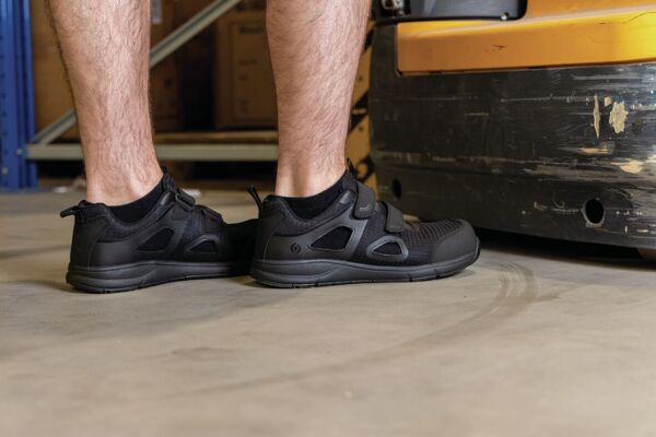 The image shows feet in black sports shoes with Velcro straps. The floor is gray and appears industrial. In the background, boxes and a yellow device can be seen.