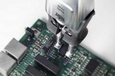 The image shows a circuit board with several black components and electrical connections. A tool for programming or testing is aimed at the board, identifiable by two gripping clamps.