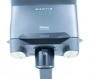 The image shows a Mantis IOTA, a compact, portable visual aid device. It has a gray surface with round lenses and a sturdy base. Ideal for improved vision for near and distant objects.