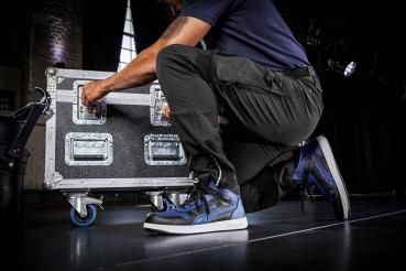 A man in a blue shirt and black pants is kneeling and opening a large, sturdy suitcase with wheels. He is wearing athletic shoes. Behind him is a stage.