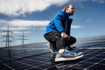 Ein Mann in blauer Jacke und sportlichen Schuhen hockt auf einer Solaranlage. Im Hintergrund sind Stromleitungen und ein klarer Himmel zu sehen. Die Szene strahlt Aktivität und Technik aus.