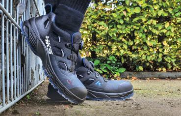 The image shows a close-up view of a black, high-cut hiking boot with blue accents, standing on a path. In the background, green plants are visible.