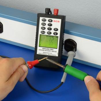 The image shows a measuring device with a screen and buttons. One hand is holding a green probe, the other a red one. These are connected by a cable that leads into the device.