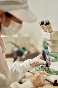 A person in protective clothing is working on a circuit board under a microscope. They are wearing gloves and a mask, and using tweezers to handle components.