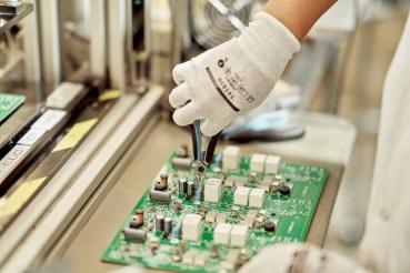 A hand in a white glove holds a pair of pliers and works on a green circuit board with various components on it. In the background, an industrial environment is visible.