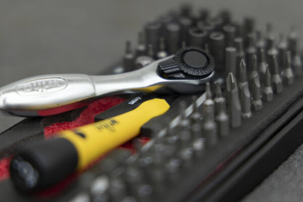 The image shows various tools. A silver ratchet lies next to a yellow screwdriver, surrounded by many black bits arranged in a red holder.