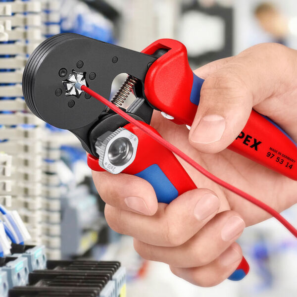 The image shows a hand holding a red and blue pair of pliers. The pliers have a black, round tip gripping a thin red cable. In the background, electrical components are visible.