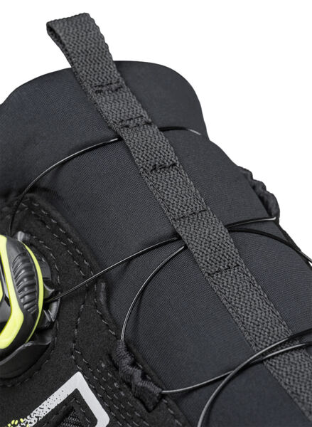 The image shows the upper part of a shoe in black with flexible laces. A round closure with yellow accents allows for easy adjustment of the fit.