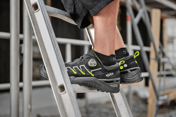 One leg stands on a rung of a ladder. The foot is wearing a solid, black shoe with green accents, which looks stable and slip-resistant. The background is blurry.