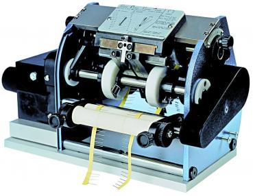 Das Bild zeigt eine mechanische Klebe- oder Etikettiermaschine. Sie hat metallische Teile, Rollen und eine Plattform, auf der ein Etikett durchläuft. Die Maschine ist robust und industriell gestaltet.