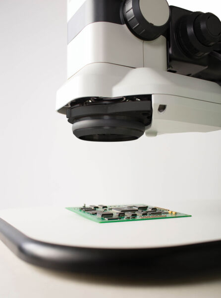 The image shows a microscope hovering over a green circuit board. The board has silver components and is situated on a light table. In the background, there is a neutral wall.