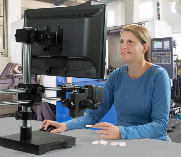 A person is sitting at a table in front of a monitor mounted on a movable stand. They are wearing a long-sleeved, blue shirt and holding small round objects in one hand.